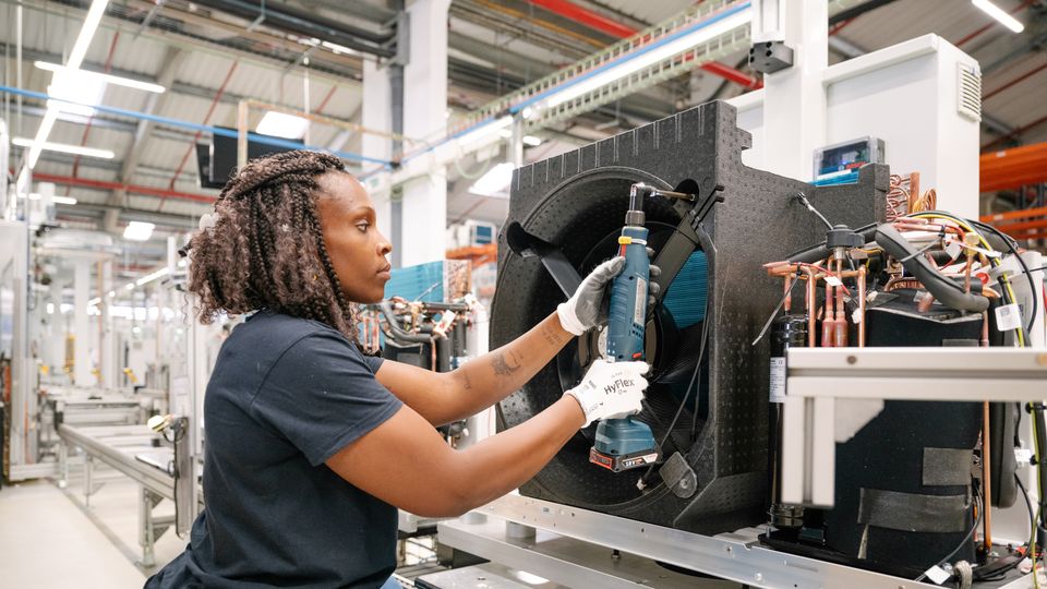 Eine Person montiert ein Bauteil an einer Wärmepumpe in einer Fabrikhalle. 